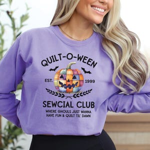 May include: A lilac sweatshirt with a Halloween-themed graphic. The design features a patchwork pumpkin, bats, and the text "QUILT-O-WEEN SEWCIAL CLUB" with the year "1999". The phrase "WHERE GHOULS JUST WANNA HAVE FUN & QUILT TIL DAWN" is also included.