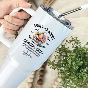 May include: White insulated tumbler with a handle and a straw. The tumbler features the text "QUILT-O-WEEN SEWCIAL CLUB" with a pumpkin graphic and the name "Sophia". The tumbler is designed for Halloween and quilting enthusiasts.
