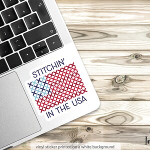 Pu&ograve; includere: Un adesivo in vinile bianco con la scritta "STITCHIN' IN THE USA" e un disegno a punto croce della bandiera americana in rosso, bianco e blu. L'adesivo &egrave; su un laptop, con uno sfondo in legno.