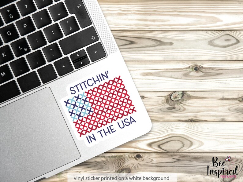 Pu&ograve; includere: Un adesivo in vinile bianco con la scritta "STITCHIN' IN THE USA" e un disegno a punto croce della bandiera americana in rosso, bianco e blu. L'adesivo &egrave; su un laptop, con uno sfondo in legno.