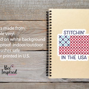 Pu&ograve; includere: Un adesivo con la scritta "STITCHIN' IN THE USA" in stile punto croce, con un disegno a bandiera rossa, bianca e blu. L'adesivo &egrave; su un taccuino a spirale, su uno sfondo di legno.