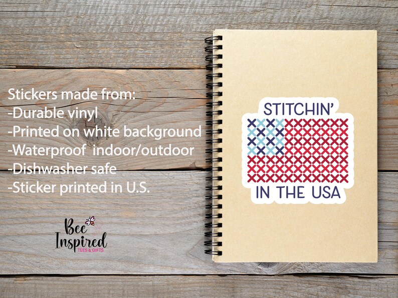 Pu&ograve; includere: Un adesivo con la scritta "STITCHIN' IN THE USA" in stile punto croce, con un disegno a bandiera rossa, bianca e blu. L'adesivo &egrave; su un taccuino a spirale, su uno sfondo di legno.