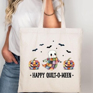 May include: Off-white tote bag featuring a Halloween-themed design. The design includes a ghost wearing a patchwork quilt, two colorful jack-o'-lanterns, and bats. The text "HAPPY QUILT-O-WEEN" is printed below the illustrations.
