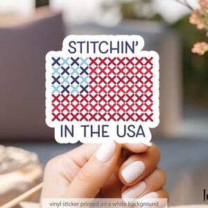 Pu&ograve; includere: Un adesivo in vinile bianco con la scritta "STITCHIN' IN THE USA" in blu navy. Il design presenta una bandiera americana a punto croce in rosso e blu. L'adesivo &egrave; tenuto in mano.