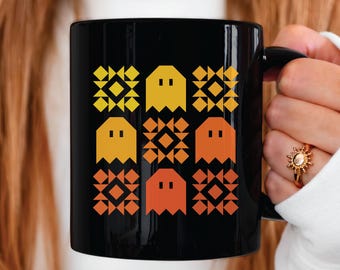 May include: Black ceramic mug featuring a repeating pattern of pixelated ghosts in shades of yellow, orange, and red. The ghosts are interspersed with geometric shapes. The mug has a black handle.