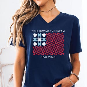May include: Navy blue V-neck t-shirt with a graphic design. The design features a flag-like pattern in red, white, and blue, with the text "STILL SEWING THE DREAM" and "1776-2026".