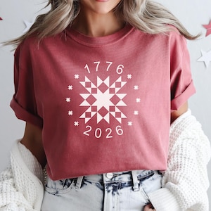 May include: Crimson-colored t-shirt with a white graphic design featuring the numbers "1776" and "2026" surrounding a starburst pattern. The shirt has rolled-up sleeves and is paired with blue jeans.