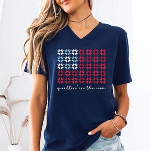 May include: Navy blue V-neck t-shirt featuring a star-patterned flag design in red, white, and blue. The text "quiltin' in the usa" is written in a cursive font below the flag. The shirt is a casual wear item.