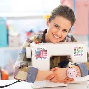 Pu&ograve; includere: Una macchina da cucire bianca con la scritta "Stitchin' in the USA" e un colorato disegno della bandiera americana. La macchina ha un cuscinetto per spilli sul lato. L'immagine &egrave; di una persona sorridente appoggiata alla macchina da cucire.
