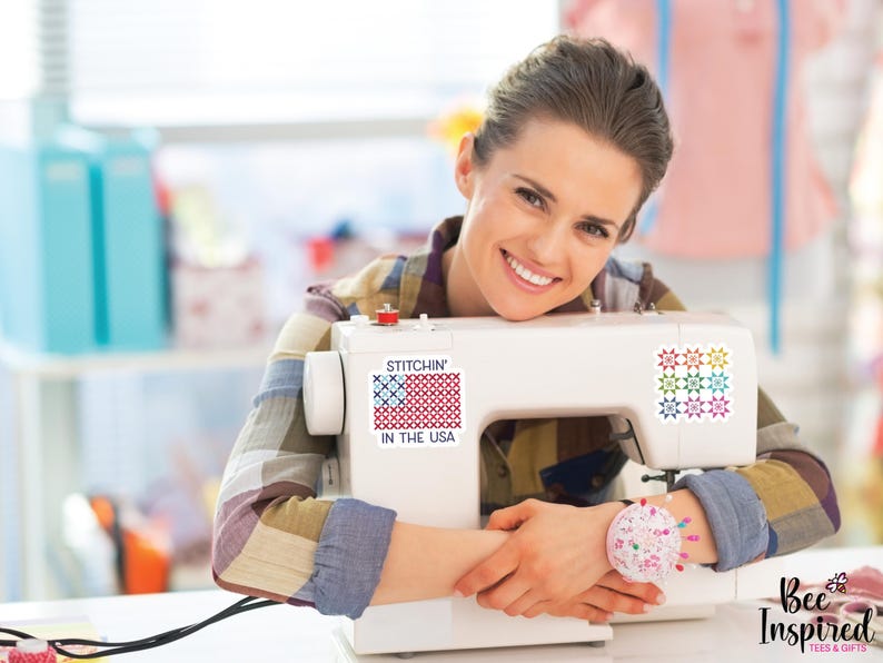 Pu&ograve; includere: Una macchina da cucire bianca con la scritta "Stitchin' in the USA" e un colorato disegno della bandiera americana. La macchina ha un cuscinetto per spilli sul lato. L'immagine &egrave; di una persona sorridente appoggiata alla macchina da cucire.