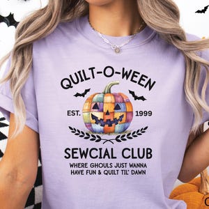 May include: A light purple t-shirt with the text "QUILT-O-WEEN" and a patchwork pumpkin graphic. Below the pumpkin is "SEWCIAL CLUB" and the phrase "WHERE GHOULS JUST WANNA HAVE FUN & QUILT TIL' DAWN".