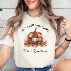 May include: Cream-colored t-shirt featuring a pumpkin design with a plaid pattern, surrounded by autumn leaves. The text reads "JUST A GIRL WHO LOVES Fall & Quilting". A fall-themed graphic tee for those who enjoy quilting.