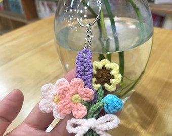 Può includere: Un portachiavi a forma di bouquet di fiori all'uncinetto con un fiocco rosa, che comprende un fiore rosa, giallo e blu, un fiore viola e un girasole marrone e giallo. Il portachiavi è attaccato a un anello portachiavi argentato.