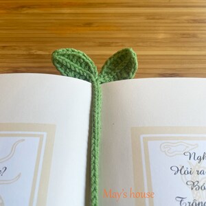 May include: A green crocheted bookmark shaped like a plant with two leaves. The bookmark is in a book with the text "May's house" visible.
