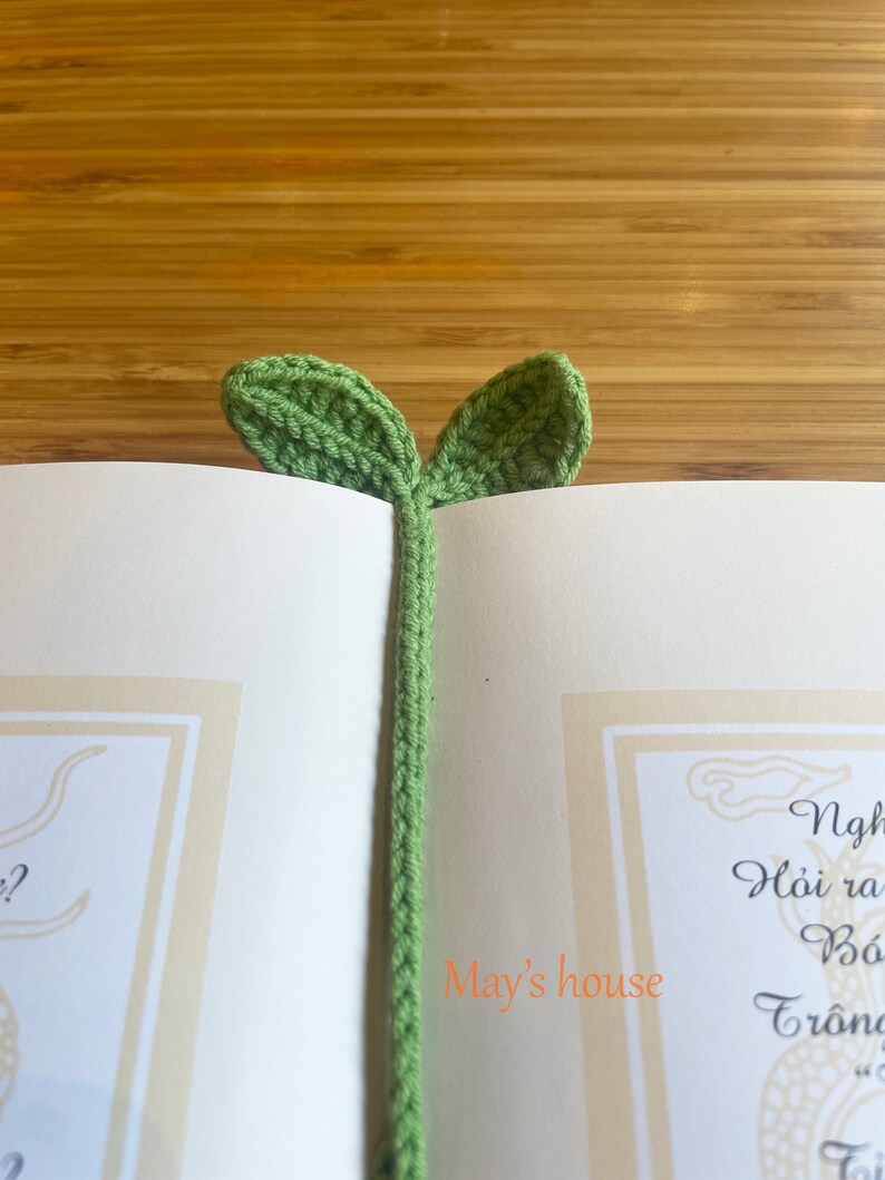 May include: A green crocheted bookmark shaped like a plant with two leaves. The bookmark is in a book with the text "May's house" visible.