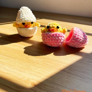 May include: Three handmade crochet chicks in eggshells. One chick is in a white eggshell, and another is in a pink eggshell. The third eggshell is pink and open. The chicks are yellow with orange beaks and black eyes. The background is a wooden surface.