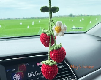 May include: A crocheted strawberry garland with a white flower hanging from the rearview mirror of a car. The car's dashboard is visible in the background. The text "May's house" is visible on the dashboard.