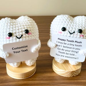 May include: Two white crochet tooth plushies with black eyes, pink cheeks, and smiling faces. One holds a sign that says "Customize Your Text." The other holds a sign that says "Happy Tooth Plush" with an inspirational message. Both are on wooden bases.
