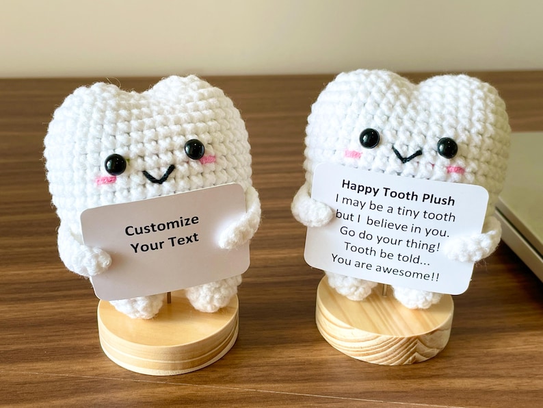 May include: Two white crochet tooth plushies with black eyes, pink cheeks, and smiling faces. One holds a sign that says "Customize Your Text." The other holds a sign that says "Happy Tooth Plush" with an inspirational message. Both are on wooden bases.