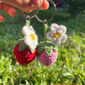 May include: Two crocheted strawberry keyrings with white flowers and green leaves attached to silver key rings. The strawberries are red and pink.