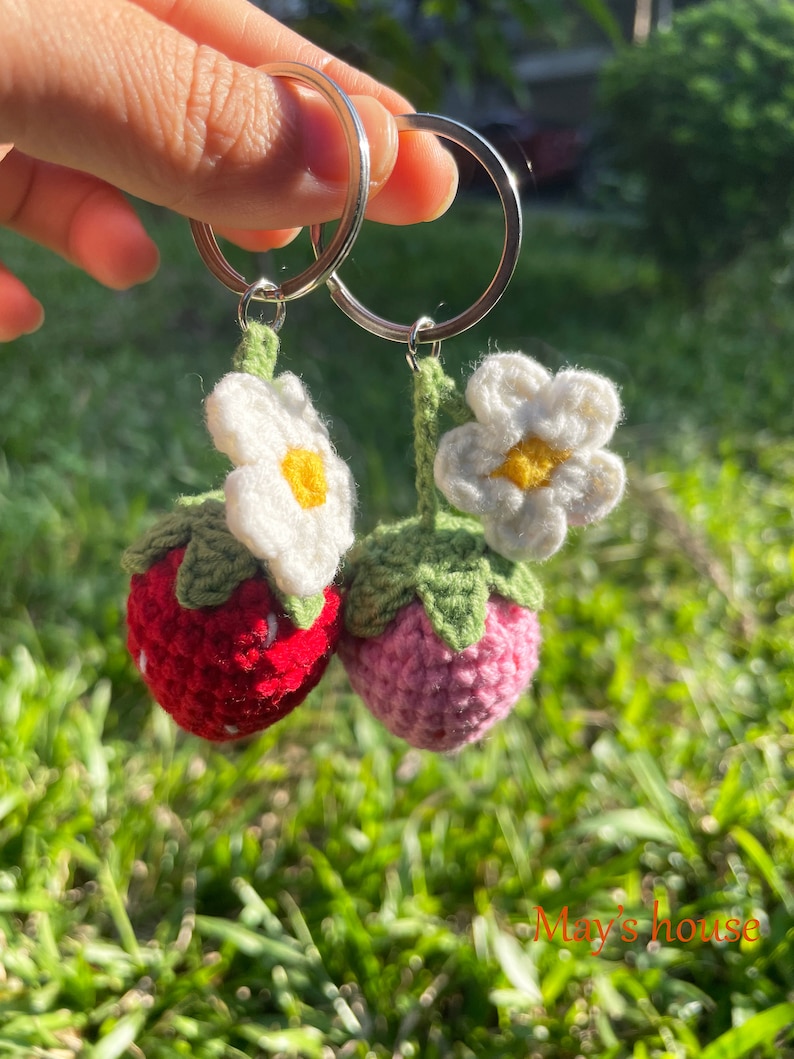 May include: Two crocheted strawberry keyrings with white flowers and green leaves attached to silver key rings. The strawberries are red and pink.