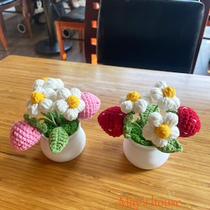 Puede incluir: Dos arreglos florales de ganchillo en macetas blancas. Cada arreglo presenta flores blancas con centros amarillos y fresas rosas y rojas.