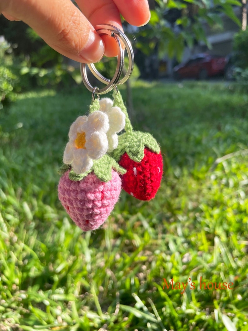 May include: A crocheted strawberry keyring with a white flower and green leaves attached to a silver key ring. The keyring is hanging from a hand.