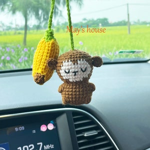 May include: A crocheted monkey with a banana hanging from its hand. The monkey is brown with white eyes and a pink nose. The banana is yellow and green. The monkey is hanging from a rearview mirror in a car. The text "May's house" is visible in the background.