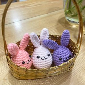 May include: Three crocheted bunny plushies in a wicker basket. The bunnies are pink, white, and purple with black eyes and embroidered smiles. The basket is gold and has a woven pattern.