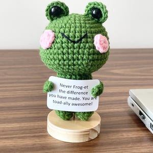 May include: A green crocheted frog figurine with black eyes and pink cheeks, standing on a round wooden base. The frog holds a white rectangular sign that reads: Never Frog-et the difference you have made. You are toad-ally awesome!
