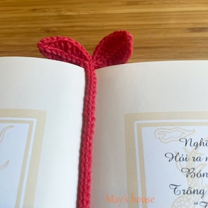 May include: A red crocheted bookmark with two leaf-shaped ends. The bookmark is on top of a book with the text "May's house" written on the page.