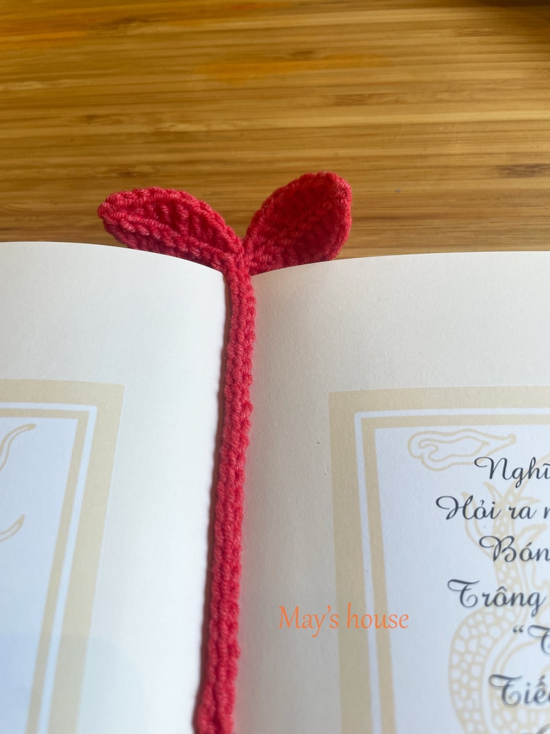 May include: A red crocheted bookmark with two leaf-shaped ends. The bookmark is on top of a book with the text "May's house" written on the page.