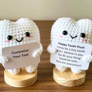 May include: Two white crochet tooth plushies with black eyes, pink cheeks, and small arms. One holds a sign that says "Customize Your Text." The other holds a sign that says "Happy Tooth Plush" with a motivational message. Both are on wooden bases.