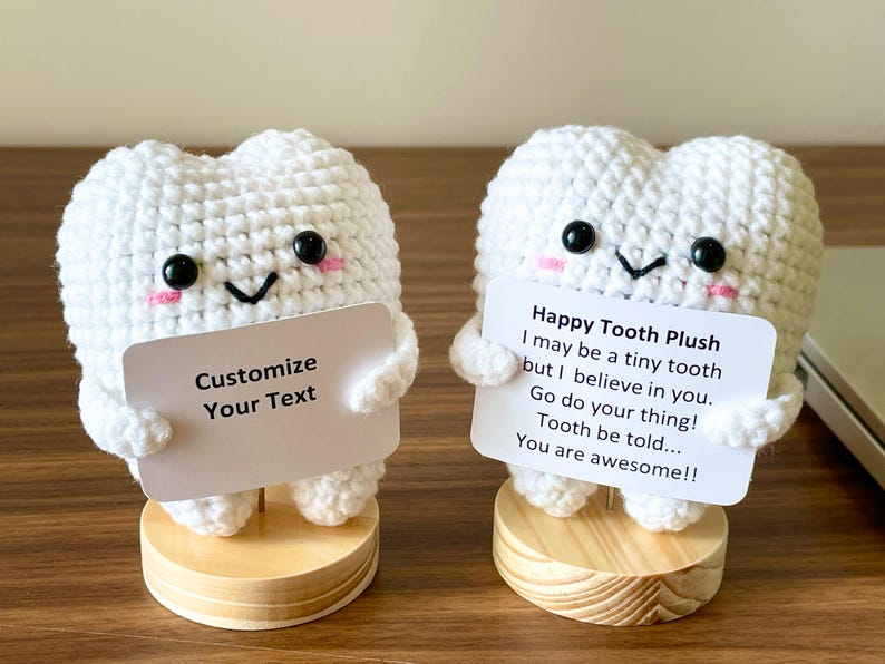 May include: Two white crochet tooth plushies with black eyes, pink cheeks, and small arms. One holds a sign that says "Customize Your Text." The other holds a sign that says "Happy Tooth Plush" with a motivational message. Both are on wooden bases.