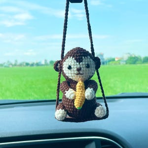 Puede incluir: Un adorno colgante de mono tejido a ganchillo en color marrón y crema, suspendido por un cordón de hilo marrón. El mono tiene una cabeza redonda, orejas pequeñas, patas blancas y sostiene un pequeño plátano amarillo hecho de hilo.