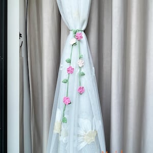 May include: White sheer curtain with embroidered floral design, held by a green crocheted tieback adorned with pink and white crocheted flowers and green leaves. The text "May's house" is visible on the lower right.