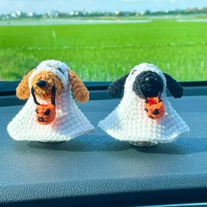 May include: Two crocheted dog figurines dressed as ghosts, one with light brown fur and one with black fur. Each dog wears a white crocheted ghost costume and holds a small orange crocheted pumpkin bucket with a black jack-o'-lantern face design.