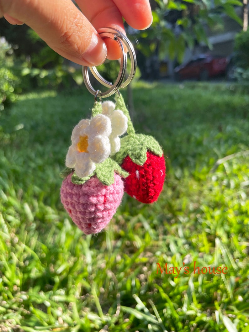 May include: A crocheted strawberry keyring with a white flower. The keyring is attached to a silver key ring.