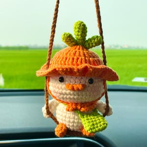 May include: A handmade crochet snowman car charm. The snowman has a carrot nose, orange hat and scarf, and a green leaf detail on top of the hat. The charm hangs from a brown rope, adding a whimsical touch to any vehicle.