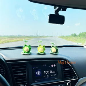 Könnte beinhalten: Drei gehäkelte Frosch-Autozubehörteile. Jeder Frosch ist grün und gelb und sitzt auf einem grünen Seerosenblatt. Das Armaturenbrett zeigt einen Radiobildschirm mit dem Text "100.0 MHz" und der Uhrzeit "9:06". Der Text "May's house" ist sichtbar.