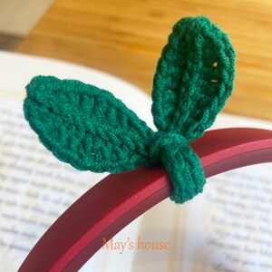 May include: A close-up of a green crocheted headband with two leaf-shaped embellishments. The headband is on a red background with the text "May's house" visible.