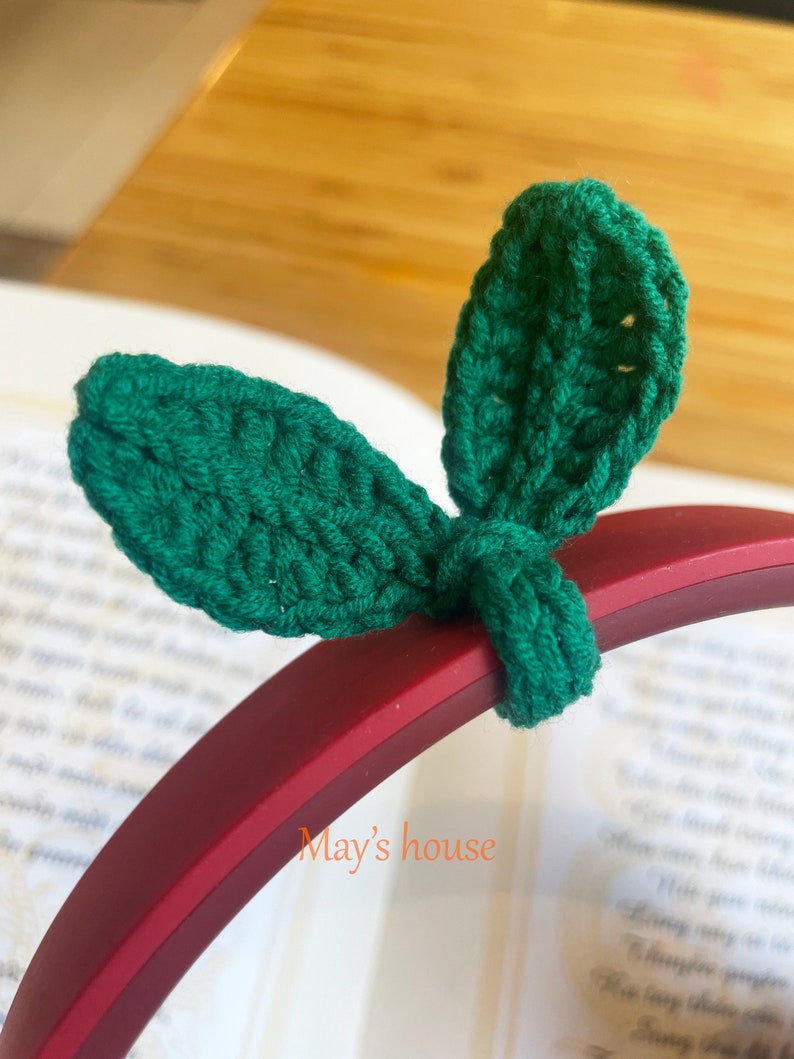 May include: A close-up of a green crocheted headband with two leaf-shaped embellishments. The headband is on a red background with the text "May's house" visible.