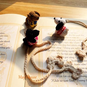 Può includere: Segnalibri a uncinetto fatti a mano a forma di cane marrone, nero e bianco. Ogni cane ha un outfit unico, con un cordoncino intrecciato abbinato. I segnalibri sono posizionati su un libro aperto. Il testo "May house" è visibile.