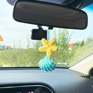 May include: A yellow crocheted starfish and a blue crocheted seashell car charm hanging from a rearview mirror. The charm is suspended by a turquoise string. The car's dashboard displays the time and radio information. The text "May's house" is visible.
