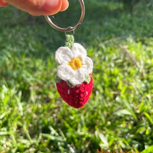 May include: A crocheted strawberry keyring with a white flower and green leaves. The keyring is attached to a silver ring.