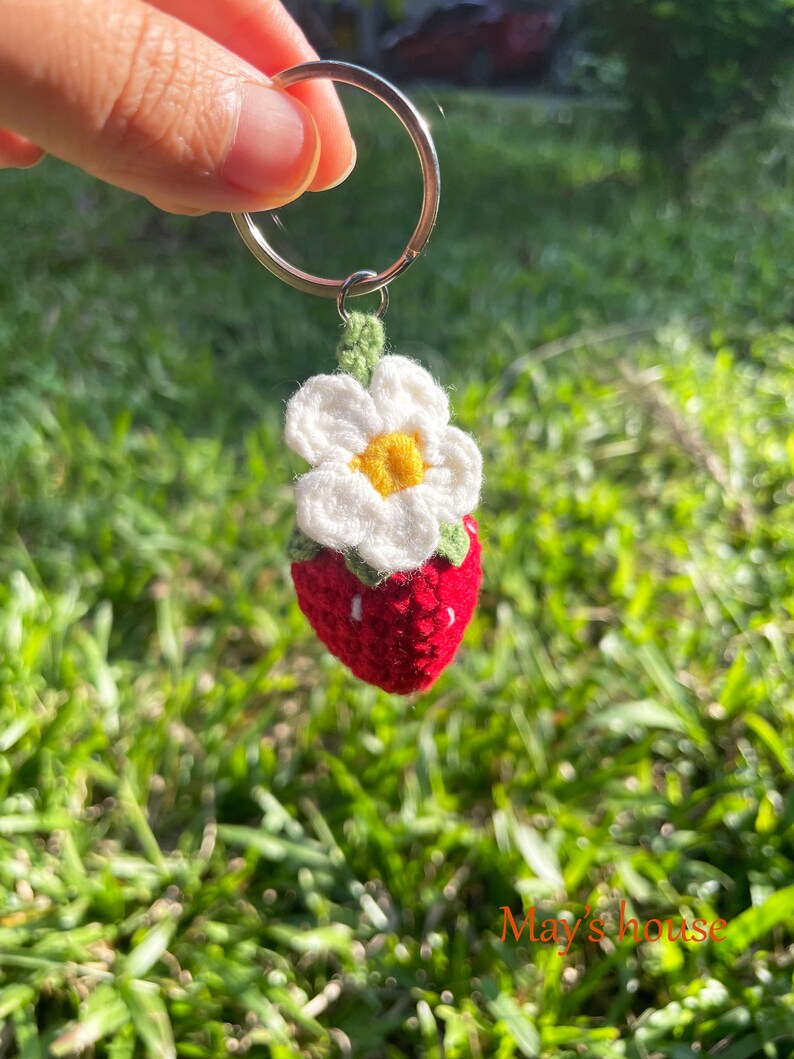 May include: A crocheted strawberry keyring with a white flower and green leaves. The keyring is attached to a silver ring.