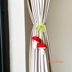 Pode incluir: Duas amarras de cortina de cogumelos vermelhos e brancos de crochê amarradas a uma cortina bege com um cordão verde. O texto "May's house" é visível no canto inferior direito.