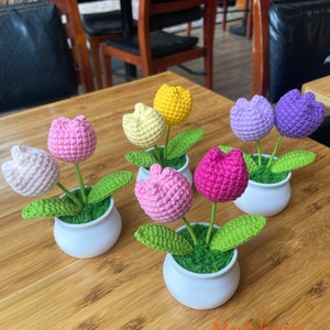 May include: Five crocheted tulip flower arrangements in white pots. The tulips are in various colors, including pink, purple, yellow, and white. The pots are sitting on a wooden table.