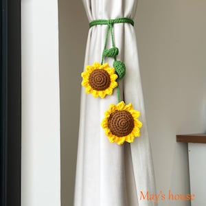 May include: Crocheted sunflower curtain tiebacks. The tiebacks feature two yellow sunflowers with brown centers and green leaves, connected by a green crocheted cord. The text "May's house" is visible in the lower right corner.