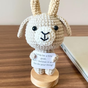 May include: A beige crocheted goat figurine with two upright horns, black bead eyes, and a white muzzle. The goat holds a small white sign that reads: I may be a tiny goat, but I believe in you. Go do your thing! It stands on a round wooden base.